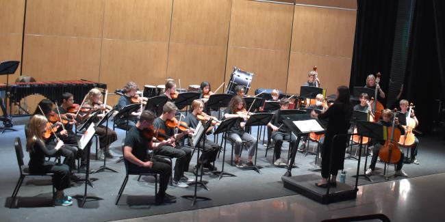 Lincoln Orchestra performing at the middle school showcase. Students seated at in chairs with their instruments as the directors cues them to play. 
