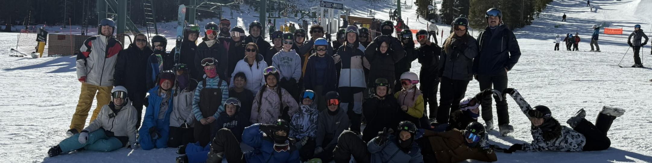 A large group of students and staff are dressed in winter gear at a ski resort where they learned how to ski or snowboard. 
