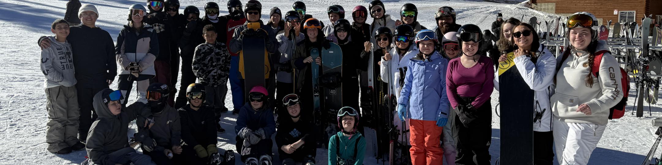 A large group of students and staff are dressed in winter gear at a ski resort where they learned how to ski or snowboard. 