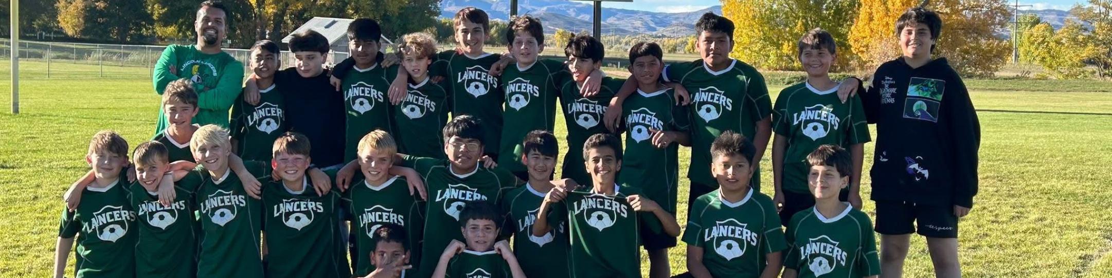 A group of middle school boys and their coach are smiling for the camera in front of the scoreboard that reads Home 4, Guests 2. They are happy because they just won the Soccer Championship for 2025.
