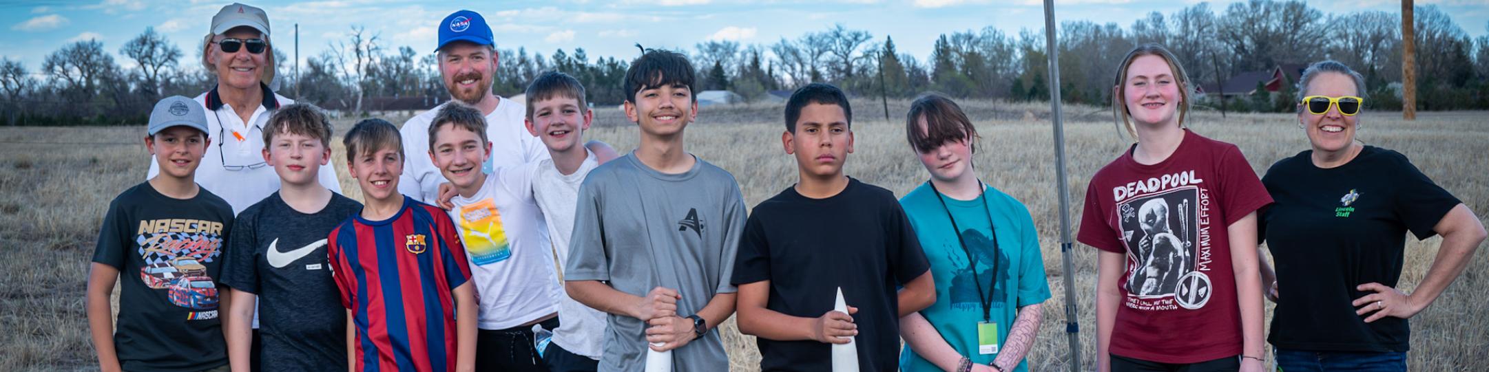 The Rocketry Club is posing for a picture with two rockets in front of them. A few adult coaches are also in the picture. It's a beautiful day, with a blue sky and clouds
