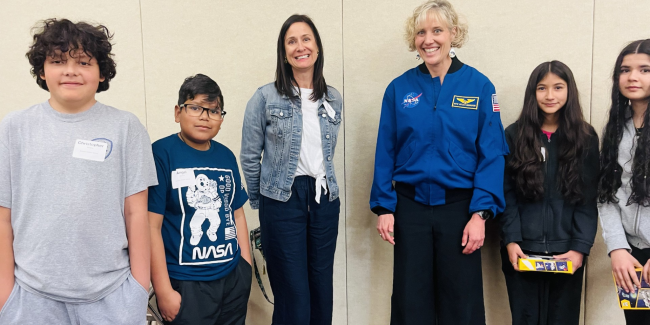 A group of kids, a teacher and an astronaut are standing in front of a yellow wall smiling.
