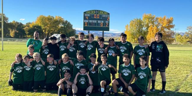 A group of middle school boys and their coach are smiling for the camera in front of the scoreboard that reads Home 4, Guests 2. They are happy because they just won the Soccer Championship for 2025.