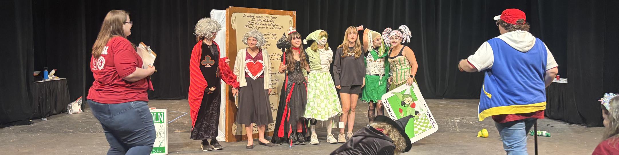 Students are on stage dressed in costumes at the Odyssey of the Mind World Finals. They have hearts and a large queen of hearts playing card. Adults are entering the stage at the end of the competition.