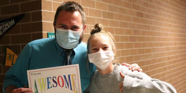 An teacher and a student are smiling. The teacher is holding a sign that says, you are awesome because