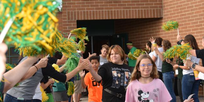 A group of middle school kids walk by adults waving green and yellow pompoms