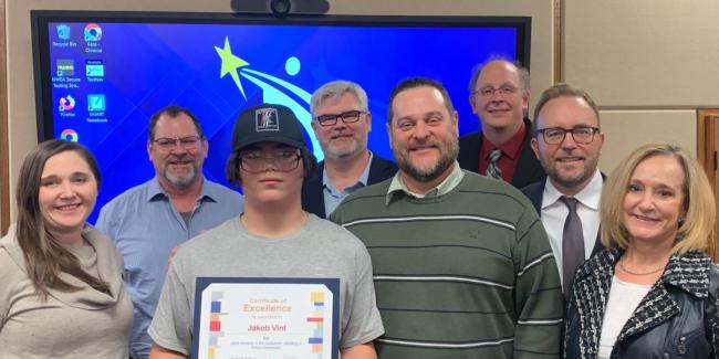 A young man with a hat on is holding an award certificate for saving a fellow student from choking. The student is surrounded by adults and all are smiling