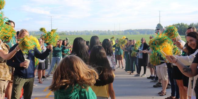 People are waving green and gold pompoms as students walk by.