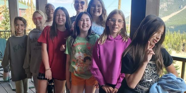 A group of middle school girls are standing on the deck of a cabin in the mountains at Camp T with smiles on their faces.