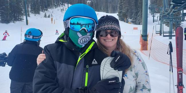 Two people are smiling at the camera. One is in snow gear with goggles and a helmet, the other person is a lady with sunglasses and a hat on. 