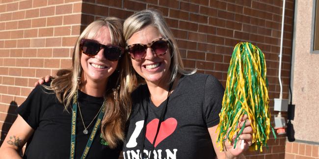 Two people are smiling at the camera, with sunglasses on and waving a green and yellow pompom.