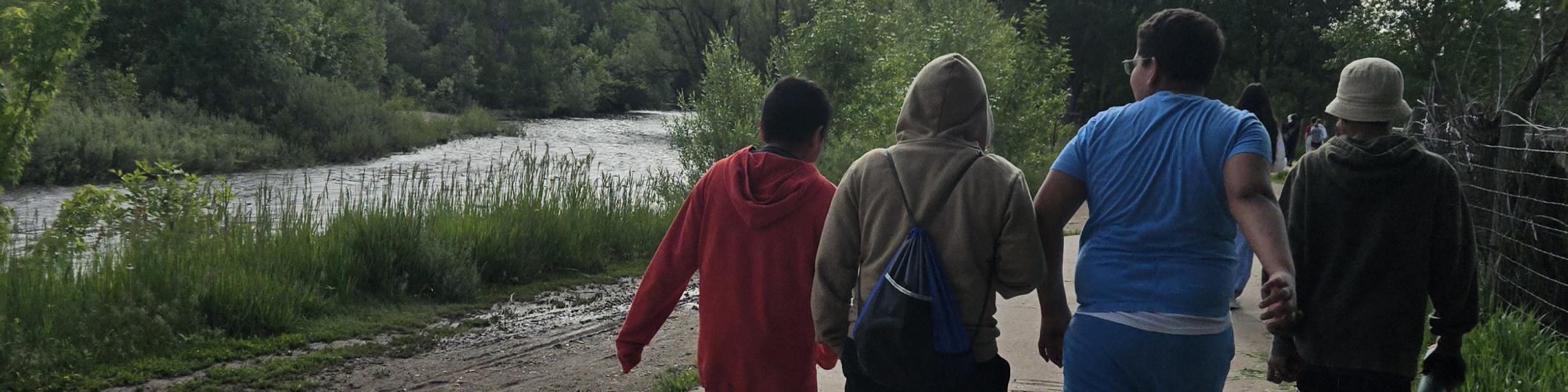 A group of middle school boys are walking along a path. A river runs next to the path. The trees are green, the sky has fluffy white clouds.