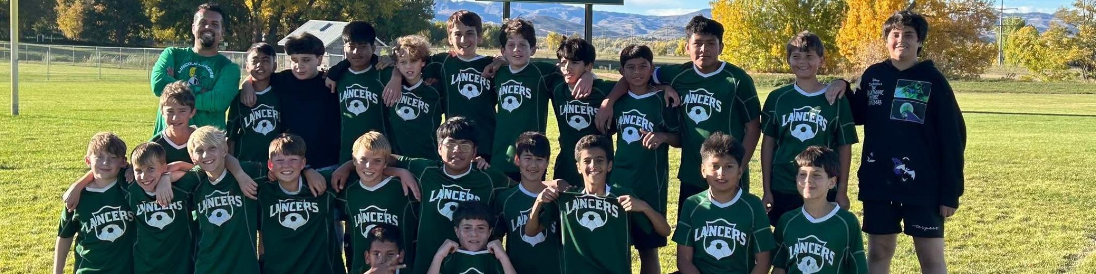 A group of middle school boys and their coach are smiling for the camera in front of the scoreboard that reads Home 4, Guests 2. They are happy because they just won the Soccer Championship for 2025.