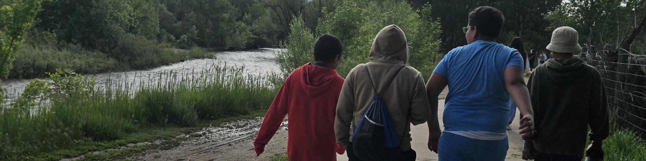 A group of middle school boys are walking along a path. A river runs next to the path. The trees are green, the sky has fluffy white clouds.