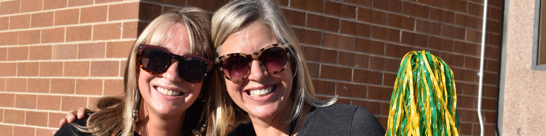 Two people are smiling at the camera, with sunglasses on and waving a green and yellow pompom.