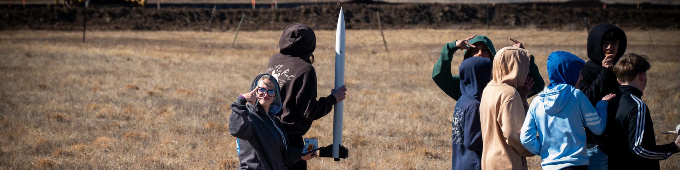 Students are in a field getting ready to launch rockets. One student is holding the rocket and one female student is looking at the camera.