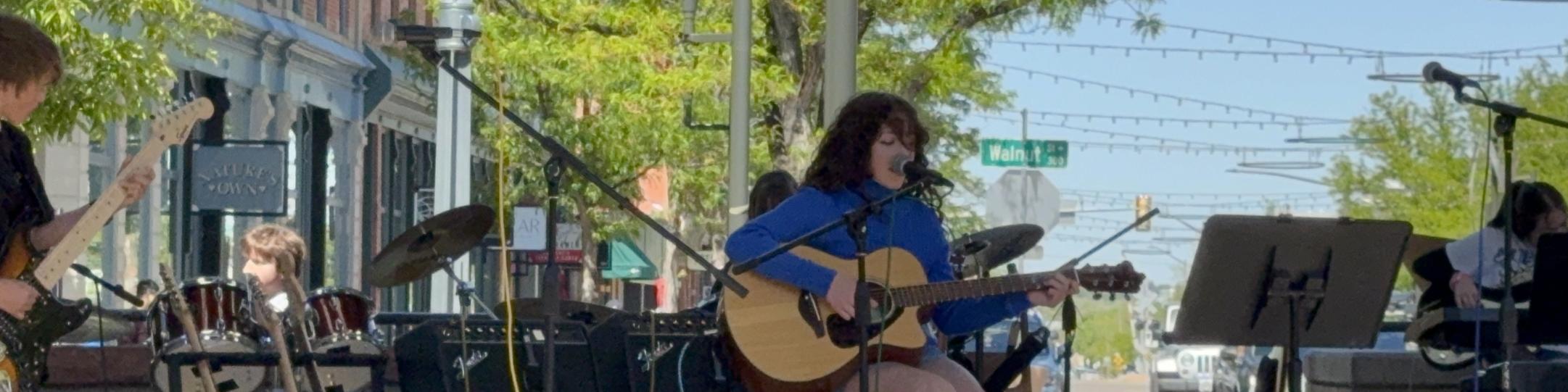 A girl in a blue top is playing the guitar with a band behind her outside on a stage. There are several students on the stage playing instruments