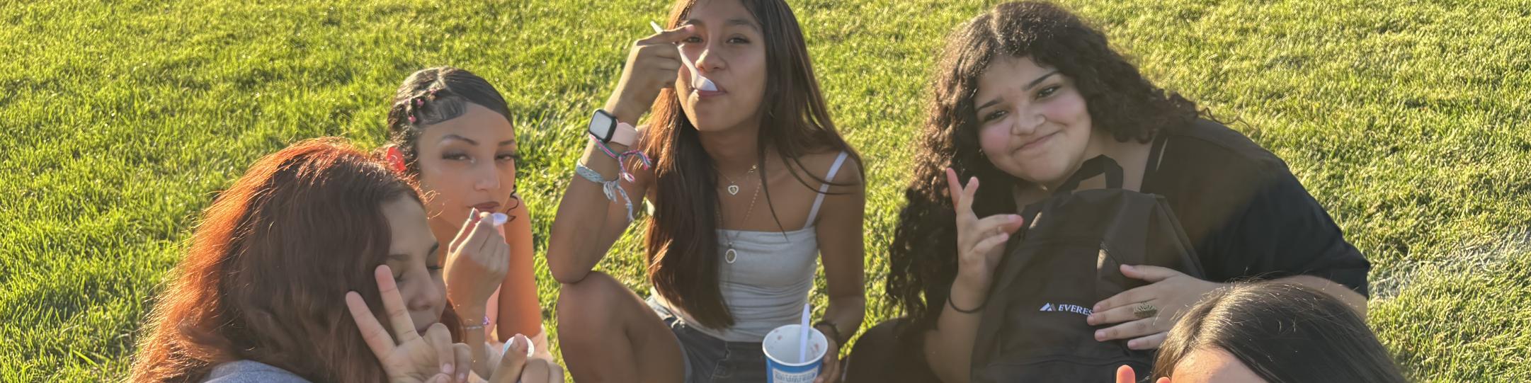 A group of girls are sitting on the green grass in the sun. They are holding up peace signs with their fingers