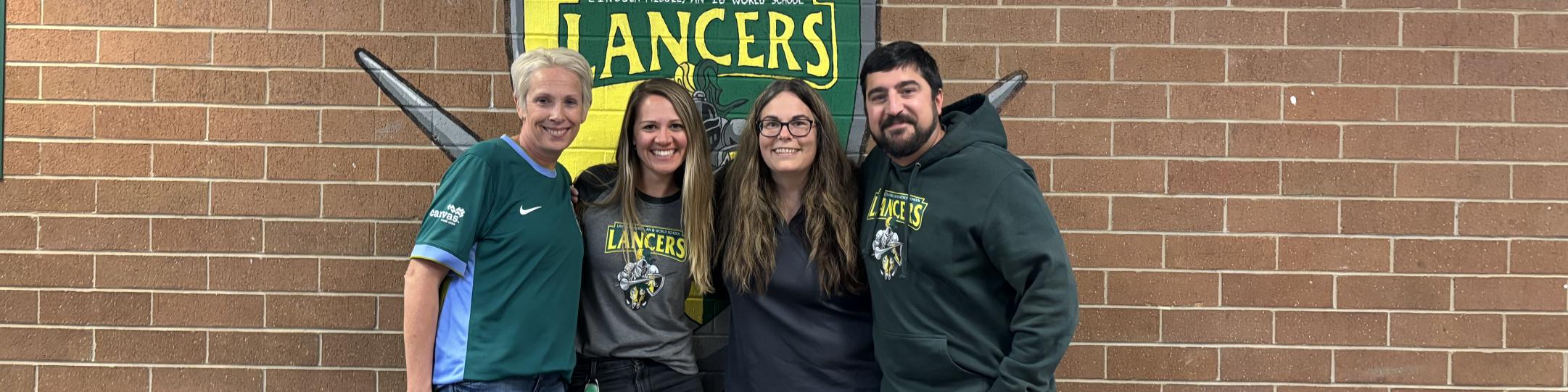 Three women and a man stand in front of signs that say Pride and Lancers. They are smiling at the camera.at the camera