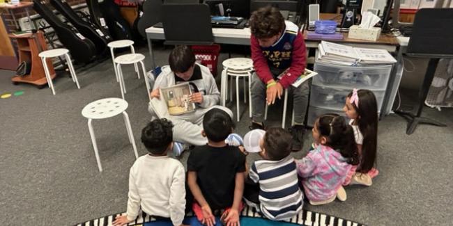 A group of elementary students are seated on a rug and middle school students are reading to them.