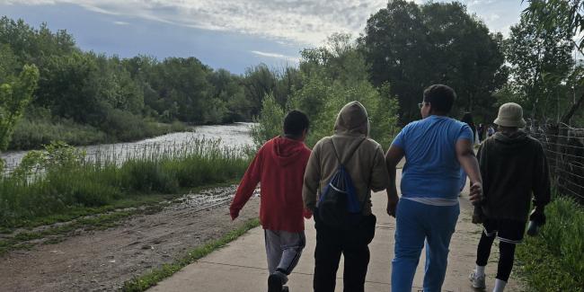 A group of middle school boys are walking along a path. A river runs next to the path. The trees are green, the sky has fluffy white clouds.
