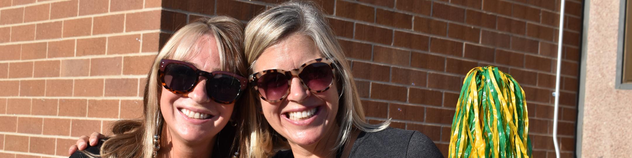 Two people are smiling at the camera, with sunglasses on and waving a green and yellow pompom.