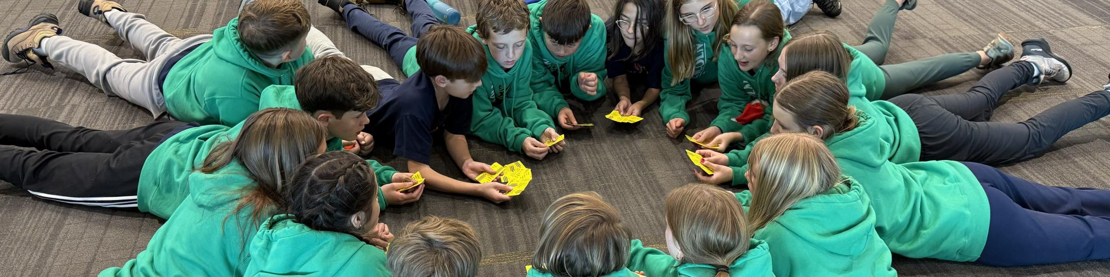 A group of students all wearing the same green sweatshirt are lying on their stomachs in a circle playing a card game. They are waiting to get onto their plane on their way to Costa Rica. 